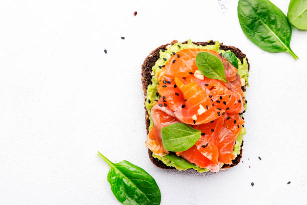 Avocado salmon sandwich or toast on rye bread with spinach, crushed cashew nuts and sesame seeds, white table background, top view stock photo