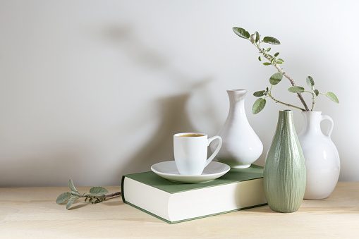 Still live of three vases with a sage leaf branch, gray green book and a coffee cup on a wooden desk or table against a light gray wall, tranquil home decoration, copy space, selected focus