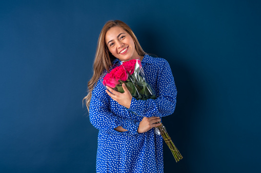 Beautiful attractive young woman posing happily in love and holding bouquet of red roses, in blue dress with dots. Isolated over blue background.