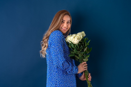 Beautiful attractive young woman posing happily in love and holding bouquet of white roses, in blue dress with dots. Isolated over blue background.