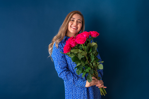 Beautiful attractive young woman posing angry with pain in her face and holding bouquet of white roses, in blue dress with dots. Isolated over blue background.