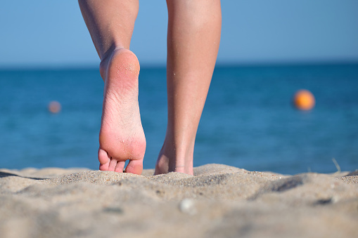 Close up of female feet walking barefoot on white grainy sand of golden beach on blue ocean water background.