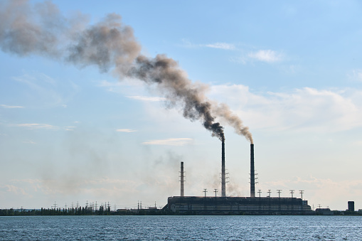 Coal power plant high pipes with black smoke moving upwards polluting atmosphere over lake water.