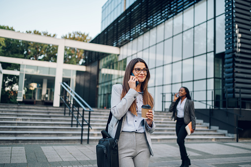 Portrait of a confident business woman wearing elegant suit walking down the street with cheerful face talking on a smartphone and holding coffee takeaway. Office building in the background.