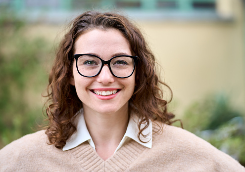 Young pretty smiling professional business woman, happy businesswoman, positive beautiful curly lady wearing glasses standing outdoor on street, looking at camera, front face headshot portrait.