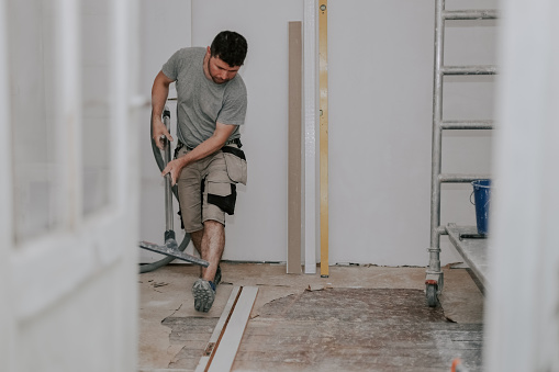 Young caucasian brunette man in work clothes on the left is vacuuming the floor with a construction vacuum cleaner in a room in an old house being renovated, side view close-up with selective focus.Construction work concept.