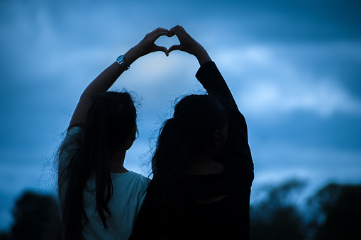 Two female best friends making heart shape symbol over blue sky.