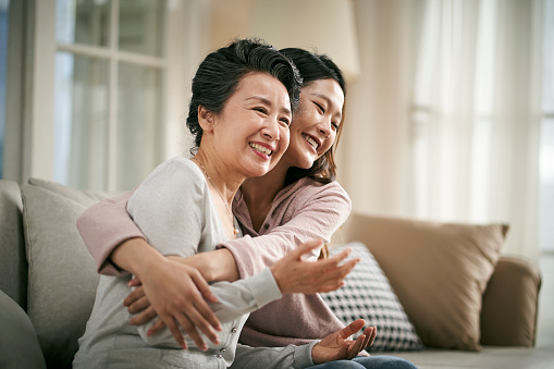 loving asian adult daughter sitting on couch at home hugging senior mother happy and smiling