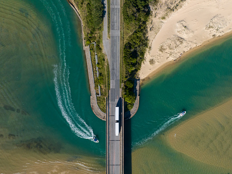 Aerial shot of a Transport truck driving along a bridge crossing a river. Two boats on a river below the container truck.
