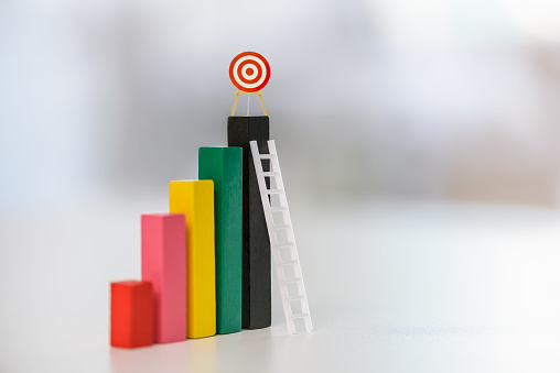 Challenging to reach a business target or attaining a company objectives, business concept : Red dartboard or bullseye perches above a black-colored wood bar graph, against which a white ladder leans.