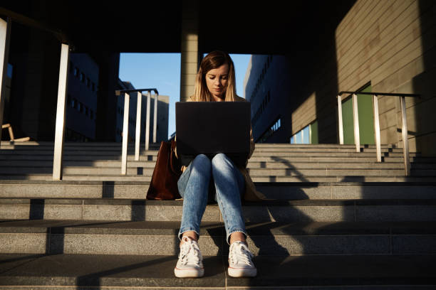 istock Woman sitting with a laptop on steps outdoors against sunset. 1455409796