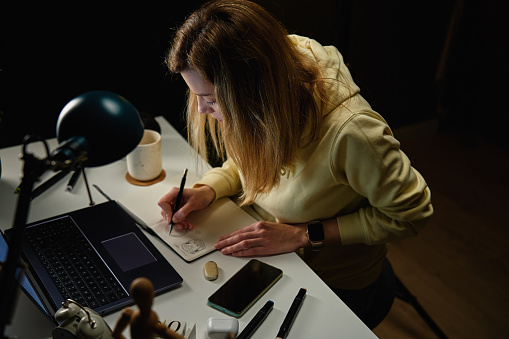 Focused woman in casual clothes looking at laptop screen and drawing illustration in notebook. Female freelancer works in home office workplace