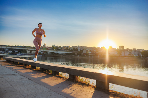 Woman in sports clothes jogging on the platform by the river at sunrise, city life workout routine