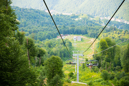 Sochi, Krasnaya Polyana, Russia - 08.10.2022: Open cable car line with people in mountains and forest on summer. Outdoor recreation in mountains, ski lift, top down view.