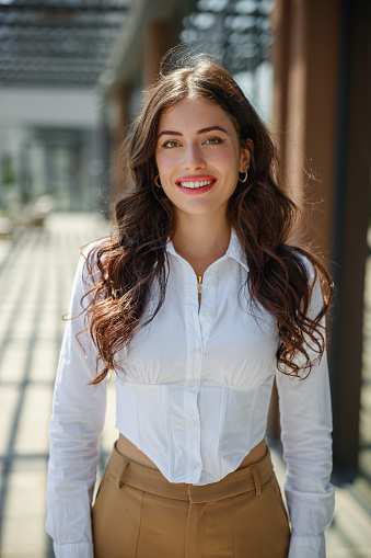 Portrait of beautiful young woman with long brown wavy hair looking at camera and smiling serenely, wearing white button down shirt and khaki pants, standing outdoors on a sunny day