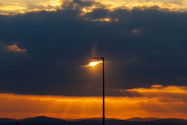 A silhouette of a light pole under a bright cloudy sunset sky