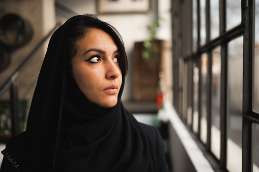 Close-up of young attractive arab woman wearing a black veil. Female profile looking out the window with serious face.