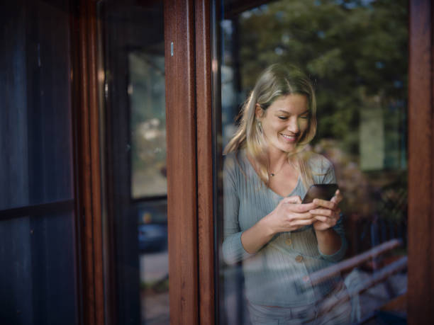 istock Happy woman using cell phone by the window at home. 1455445528