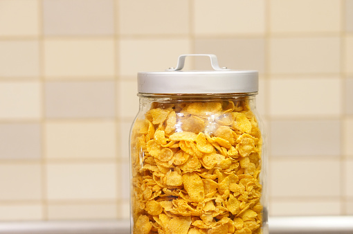A closeup of corn flakes in a glass container against a tile wall