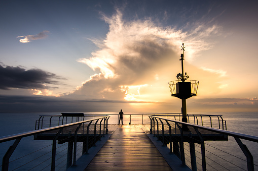 A person standing on the bridge over the calm sea under a cloudy sky at sunset in Spain