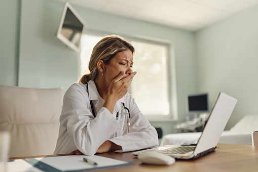 Tired medical expert yawning while reading an e-mail on a computer in the office at hospital.