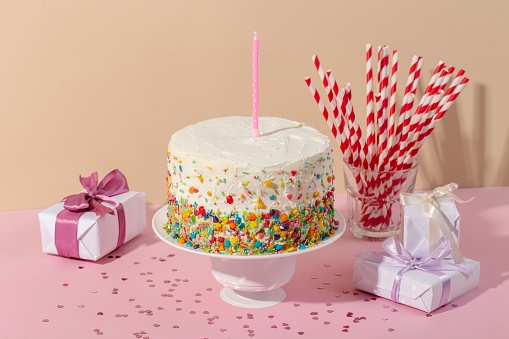 A birthday cake with colorful candies and  pink candle on it and gift boxes on a purple background