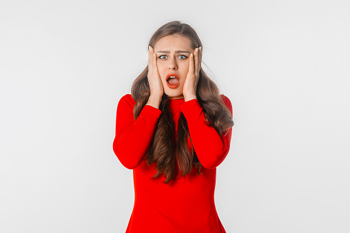 Amazed young woman. Portrait of beautiful brunette girl with shocked facial expression keeps hands on the face, standing with open mouth against white studio background