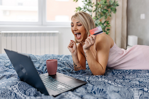 A young Caucasian woman is cheerfully clenching her fists and smiling wide, while holding a credit card and looking at her laptop.