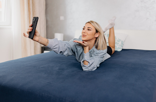 A young Caucasian woman is lying on her stomach on a bed, filming herself cheerfully blowing a kiss.