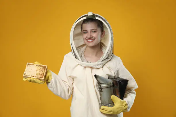 Beekeeper in uniform holding smokepot and hive frame with honeycomb on yellow background