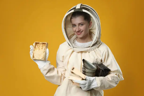 Beekeeper in uniform holding smokepot and hive frame with honeycomb on yellow background