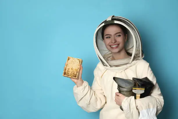 Beekeeper in uniform holding smokepot and hive frame with honeycomb on light blue background. Space for text