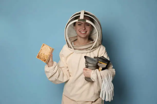 Beekeeper in uniform holding smokepot and hive frame with honeycomb on light blue background