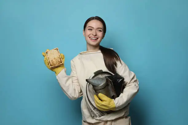 Beekeeper in uniform holding smokepot and hive frame with honeycomb on light blue background