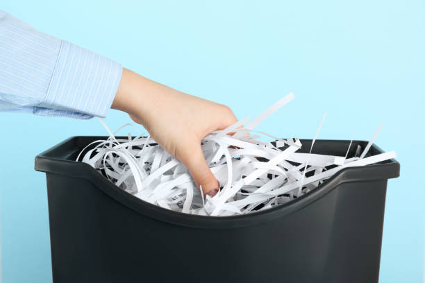 istock Woman putting shredded paper strips into trash bin on light blue background, closeup 1455468119