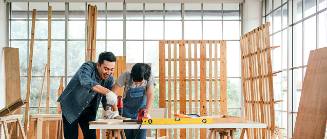 Asian father and son work as a woodworker or carpenter, Father teaches his son to drill holes in a wooden plank carefully together with teamwork. Craftsman carpentry working at home workshop studio.