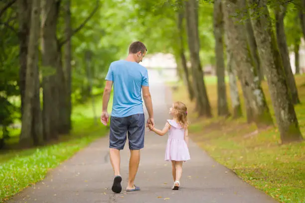 Photo of Little daughter and young adult father speaking and walking on sidewalk through tree alley at city park. Spending time together in beautiful warm sunny summer day. Back view.