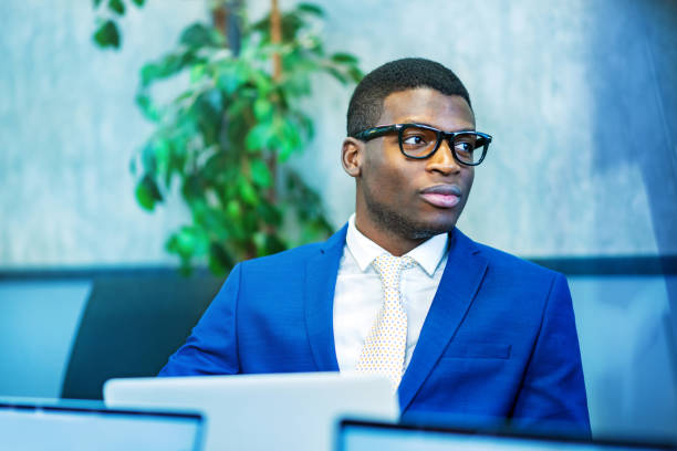 African American male entrepreneur in blue suit and stylish glasses looking away while using laptop during work in modern office