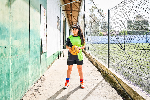 Full length portrait of soccer teenage girl looking at camera holding a ball. Confident female player wearing green jersey on sports field.