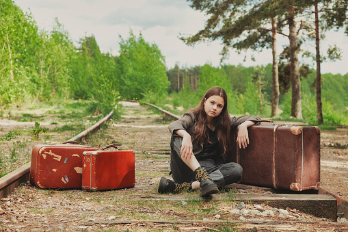 Teen girl in stylish retro image with vintage suitcases sitting on railway in summer woodland, looking at camera. Fashionable young lady on forest railroad. Travel vacation concept. Copy text space