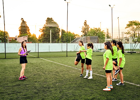 Female coach explaining game strategy to girls soccer team on sports field. Mature woman instructor sharing a game plan with female players on playground.
