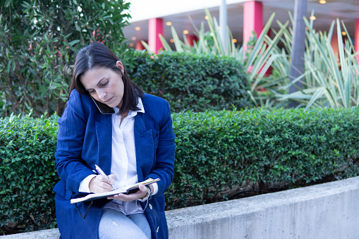 Beautiful Caucasian businesswoman calling phone and wrting in a notebook outside an office building. She is wearing a casual blue jacket, copy space