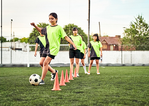 Girls soccer players training on playground. Teenage girl kicking a ball around cones on sports field with other children waiting their turn to do the drill.