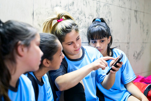 Girls soccer team using mobile phone after soccer practice. Female sports team sitting in locker room after training session.