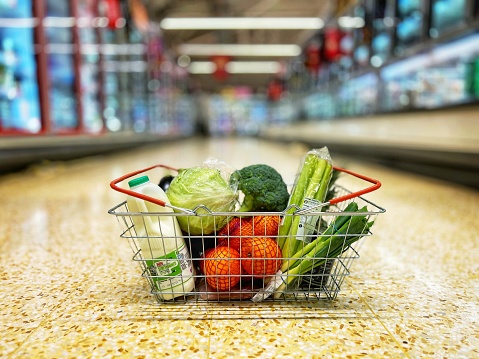Close up of a shopping basket full of healthy food, including an assortment of fruit and vegetables, on the floor of a supermarket aisle. The products on the shelves are defocused in the background.