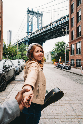 A redhead woman is holding the hands of her husband, telling him to follow her. Manhattan Bridge in the background.