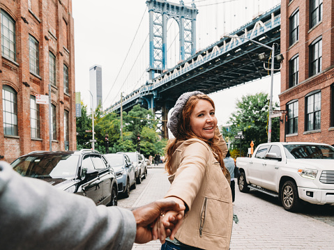 A redhead woman is holding the hands of her husband, telling him to follow her. Manhattan Bridge in the background.