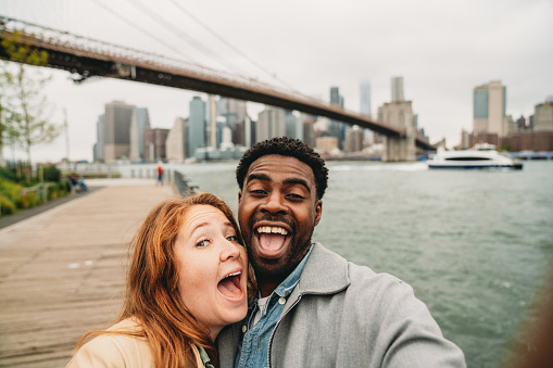 A multi-ethnic couple is taking a selfie in New York City with the skyline in the background. The are bonding together. Brooklyn Bridge in the background.