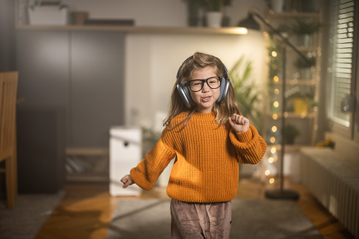 Portrait of a beautiful little girl listening music on headphones while standing in living room at home.