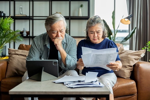 old retired asian senior couple checking and calculate financial billing together on sofa involved in financial paperwork, paying taxes online using e-banking laptop at living room home background